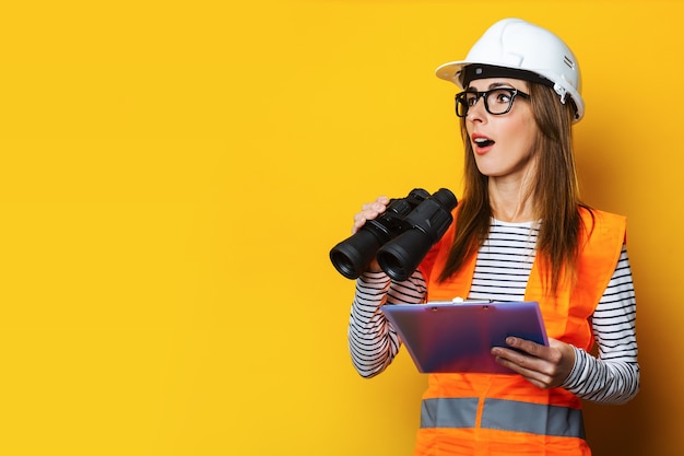 La giovane donna con una faccia sorpresa in una maglia e un cappello duro tiene una lavagna per appunti e un binocolo su colore giallo