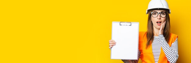 Young woman with a surprised face in a vest and hard hat holds a clipboard on a yellow