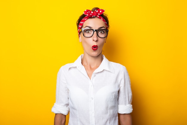 Young woman with a surprised face, wearing glasses and a headband on her head on a yellow wall.