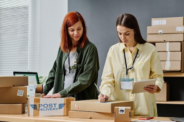 Young woman with tablet writing down address of receiver on packed box