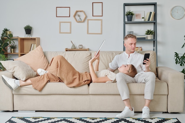 Young woman with touchpad keeping her head on knees of husband using smartphone while lying on couch and scrolling through online pages