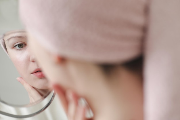 Young woman with a towel on head apply cream on face skin and doing makeup at home