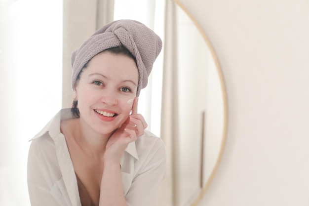 молодая женщина с полотенцем на голове смотрит в зеркало и наносит крем на кожу лица