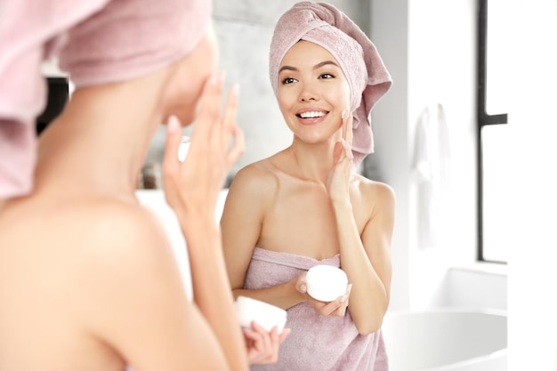 Young woman with towels applying facial cream near mirror in bathroom