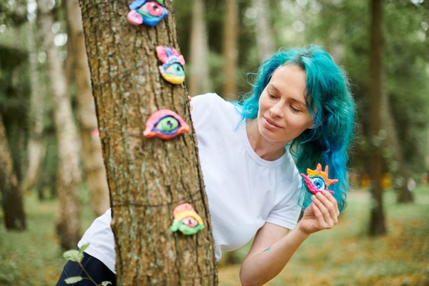 Young woman with turquoise dyed hair in white T shirt considering handmade colorful eye amulet