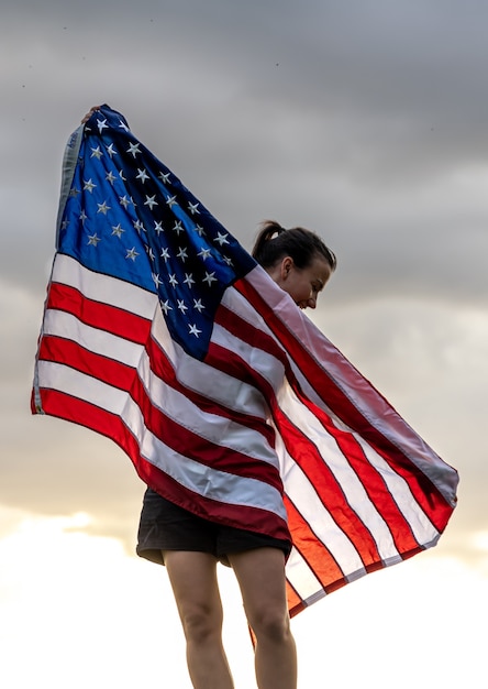 写真 自然の中で夕日を楽しんでいる米国旗を持つ若い女性。
