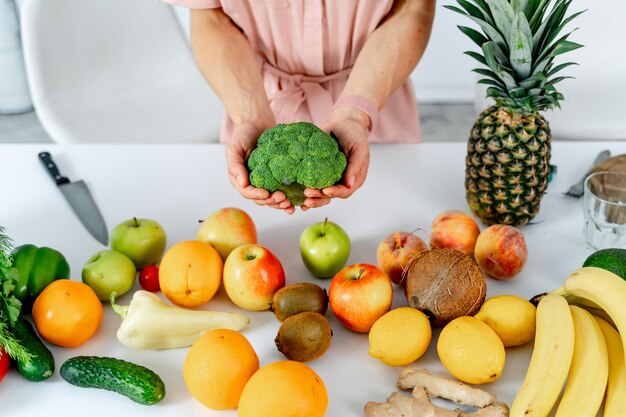 ブロッコリーを手に持ってキッチンで調理する野菜を持つ若い女性。健康食品のテーブル。トリミングされた写真。