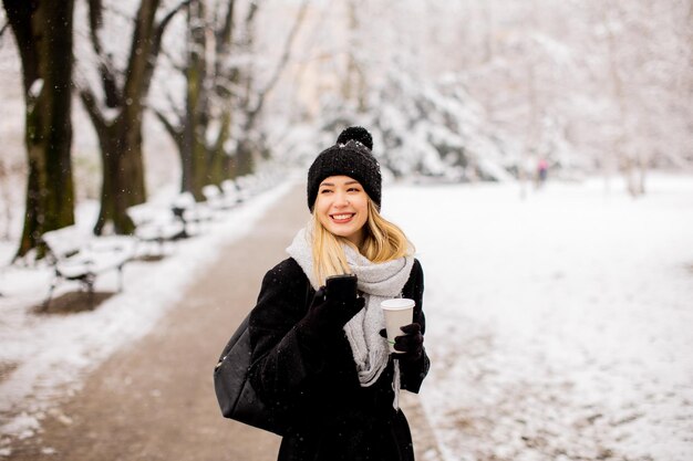 行くコーヒーを飲みながら寒い冬の雪の中で暖かい服を着た若い女性