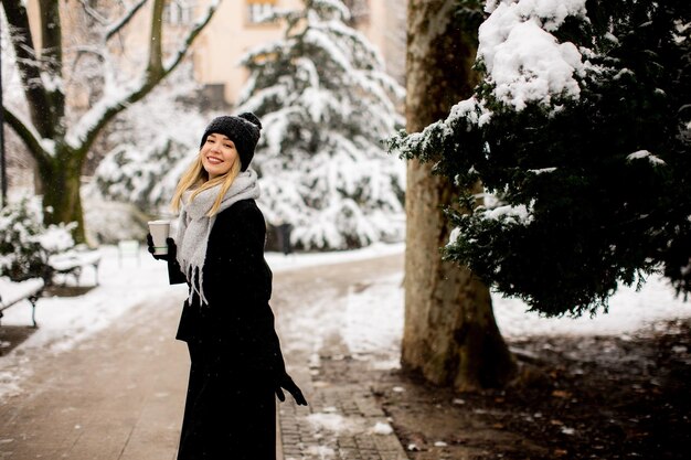 行くコーヒーを飲みながら寒い冬の雪の中で暖かい服を着た若い女性