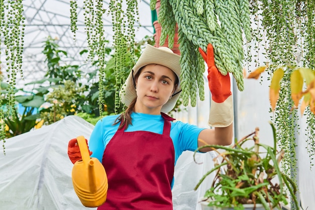 Молодая женщина с лейкой работает в теплице среди ампельных растений