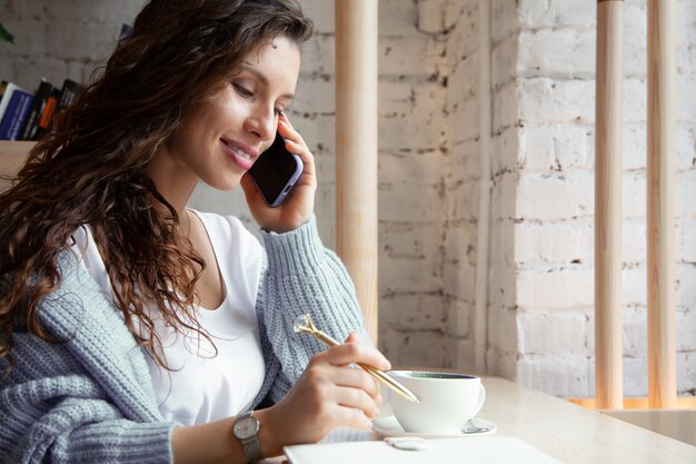 Young woman with wavy hair in warm blue knitted sweater is talking on phone and writing notes while drinking healthy hot blue coffee latte made from natural butterfly pea tea. Beauty and wellbeing