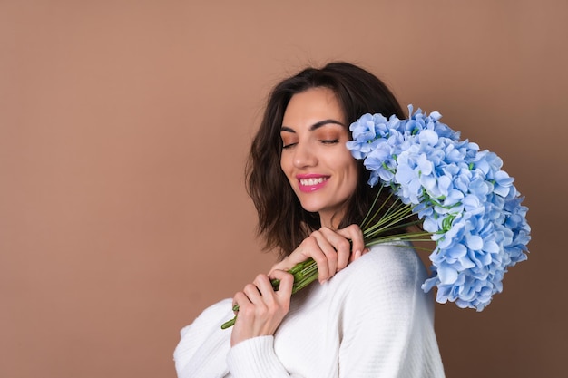 Una giovane donna con capelli ondulati e voluminosi su uno sfondo beige con un rossetto rosa brillante e un lucidalabbra in un maglione bianco tiene un mazzo di fiori blu