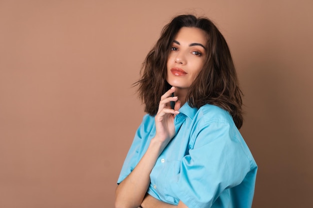 Young woman with wavy voluminous hair and natural daytime makeup wearing a blue shirt on a beige background