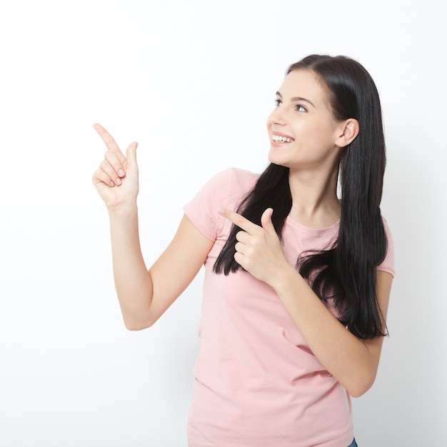 Молодая женщина с белой идеальной улыбкой в летней одежде показывает указательные пальцы в сторону на пустом месте над белой стеной.