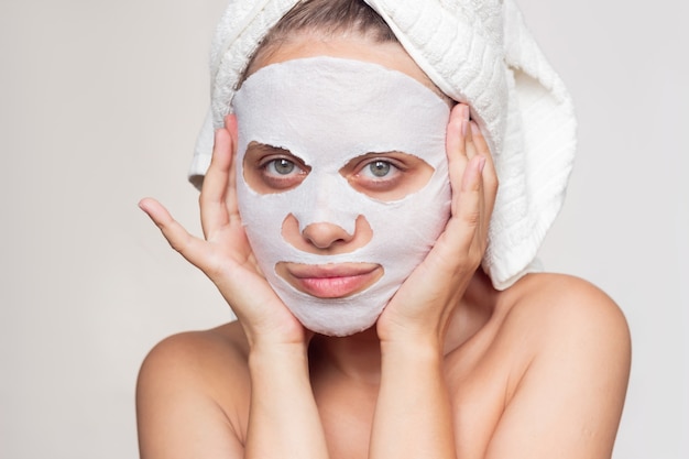 A young woman with a white towel on head puts a fabric cosmetic moisturizing mask on her face