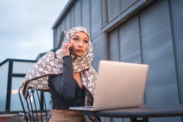 young woman with a white veil at the computer on the terrace of a coffeee shop, with the computer and talking on the phone.