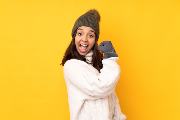 Молодая женщина в зимней шапке на изолированном желтом фоне празднует победу