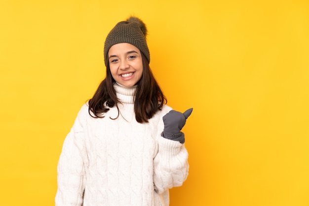 Giovane donna con cappello invernale su giallo isolato che punta di lato per presentare un prodotto