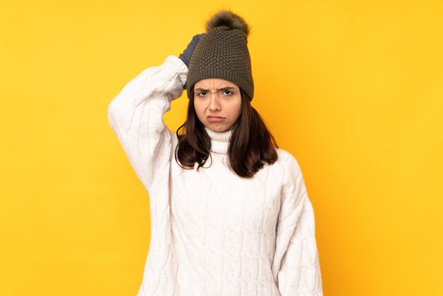 Young woman with winter hat over isolated yellow wall having doubts while scratching head