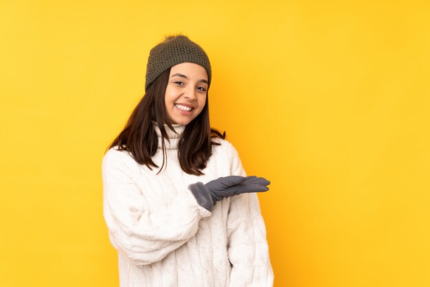 Young woman with winter hat over isolated yellow wall presenting an idea while looking smiling towards