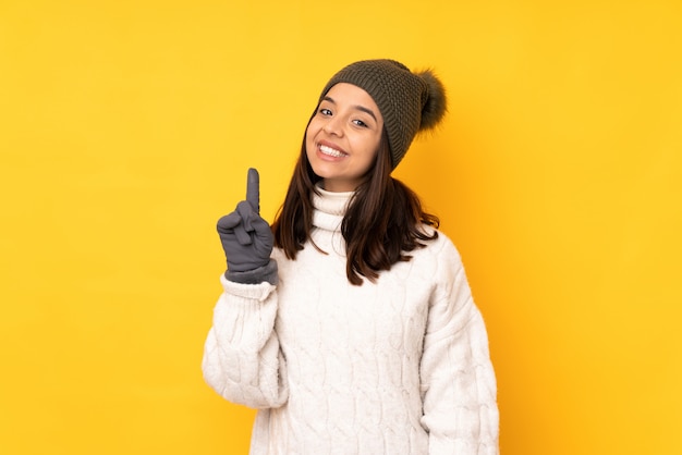 Young woman with winter hat over isolated yellow wall showing and lifting a finger in sign of the best