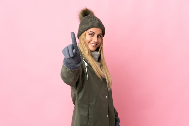 Giovane donna con cappello invernale sulla rappresentazione rosa e alzando un dito