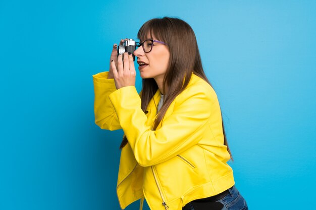 Young woman with yellow jacket on blue wall holding a camera