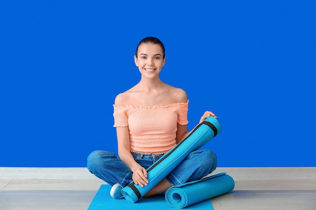 Young woman with yoga mats sitting near color wall