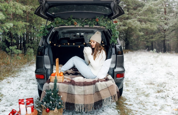 A young woman in a wool hat sits in the trunk of a car and holds a Cup of hot tea