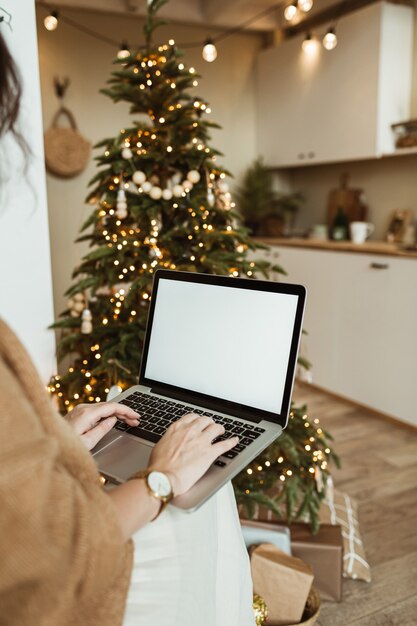 Young woman work on laptop computer with blank display screen with copy space. Modern home living room interior design decorated for Christmas celebration with Christmas tree.