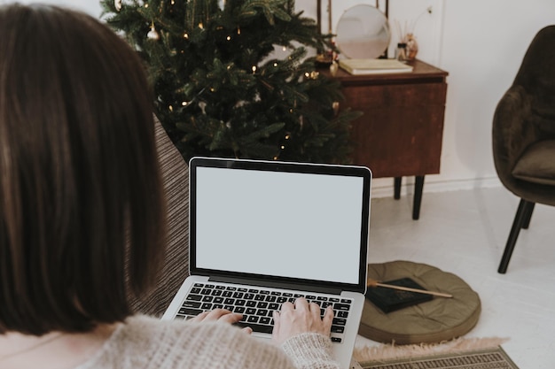 Young woman work on laptop computer with blank screen with mockup copy space Modern home living room interior design decorated for Christmas New Year celebration with Christmas tree