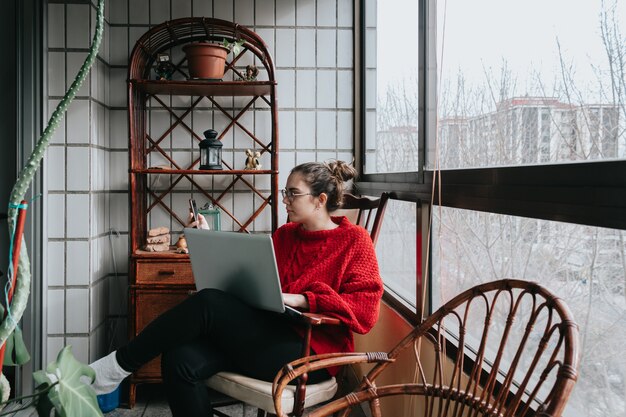 A young woman working on his laptop sitting on a wooden chair on a crystal gallery during a bright day in the city