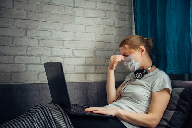 Young woman working at home during quarantine. Sick woman wearing face mask with laptop, lying on sofa. Remote office, self-isolation concept