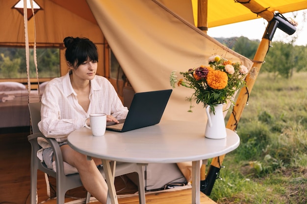 Молодая женщина работает на ноутбуке, сидя возле большой палатки на природе