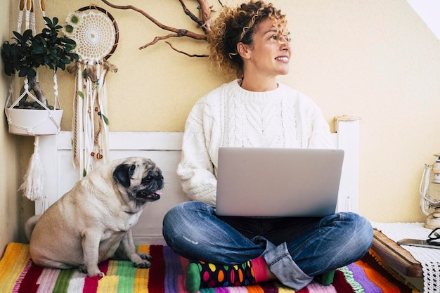 Молодая женщина работает на ноутбуке со своей лучшей подругой собакой, сидящей рядом с ней на скамейке дома. Образ жизни владельца собаки-мопса. Люди вместе с животным. Удаленная новая нормальная современная умная работа.
