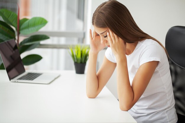 Young woman working at office desk in front of laptop suffering from chronic daily headaches