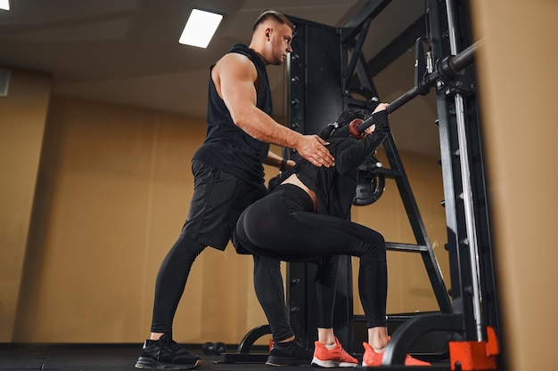 Young woman working out legs with barbell in a gym with instructor squat exercise personal trainer