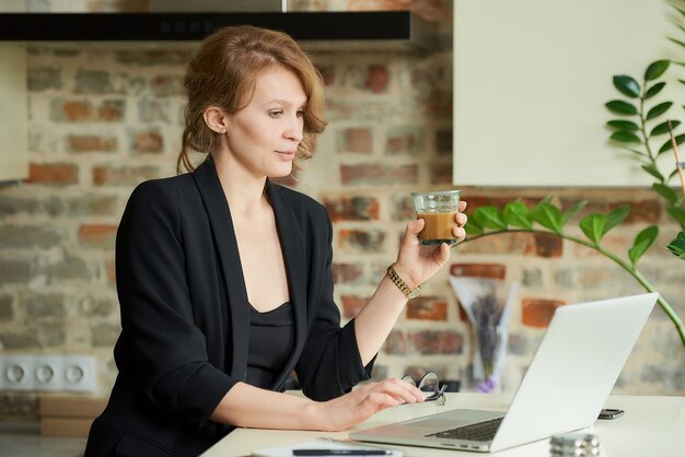 A young woman working remotely in her kitchen. A female boss with a coffee listening to a discussion of a project with employees at a video conference at home.