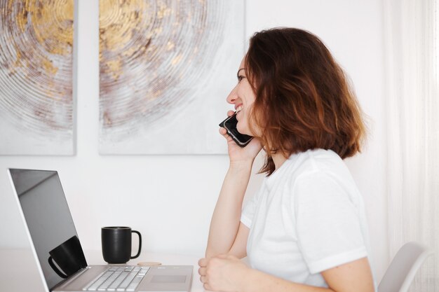 Young woman working with computer at home and talking on smartphone. Cheerful, positive girl works in home office. Work with laptop, white and modern interior.