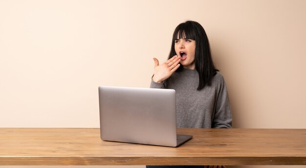 Young woman working with her laptop yawning and covering wide open mouth with hand