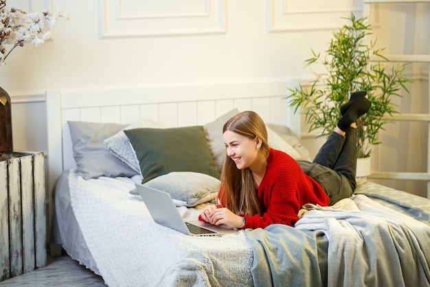 La giovane donna lavora al computer, seduta su un letto, lavorando a distanza. una ragazza con i capelli lunghi in un maglione rosso e jeans lavora a casa.