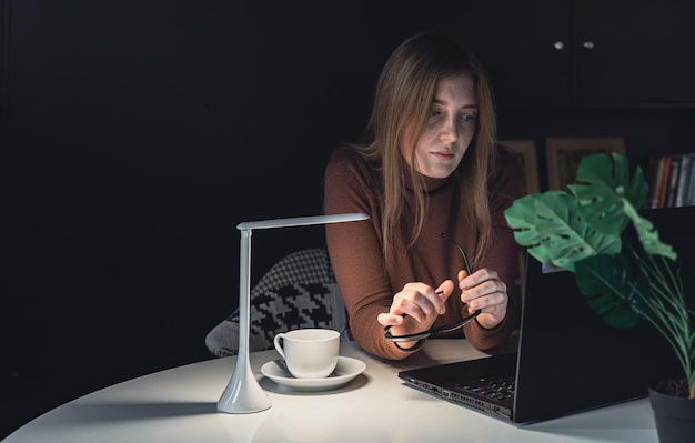 A young woman works behind a laptop at night with a lamp light