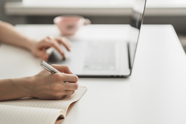 Young woman works on a laptop and takes notes in a notebook 