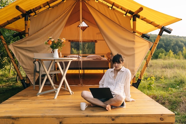 Young woman works on laptop while sitting relaxed on chair by the campfire traveling with tent Concept of remote work and escape to nature