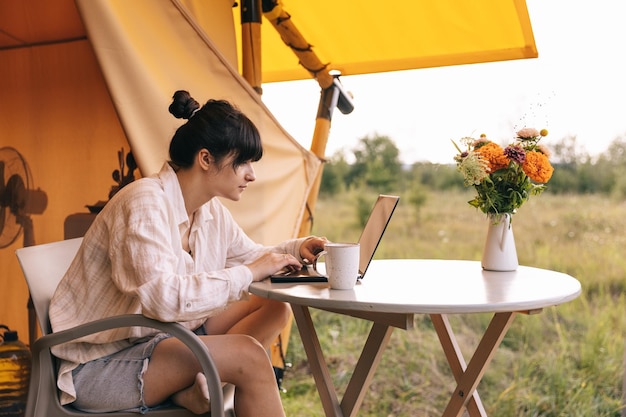 Young woman works on notebook while sitting relaxed on chair by the campfire traveling with tent Concept of remote work and escape to nature