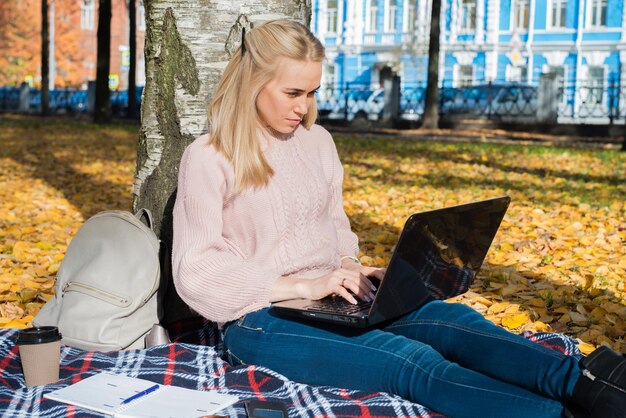 A young woman works in a park typing on a laptop