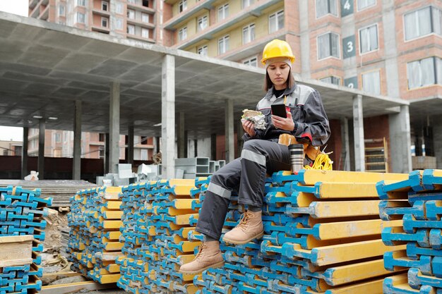 Young woman in workwear and protective helmet scrolling in smartphone