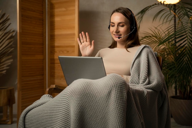 Young woman wrapped in a blanket with a headset uses a laptop computer accepts video calls sits on