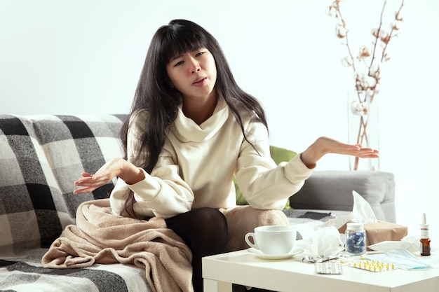 Young woman wrapped in plaid looks sick ill sneezing and coughing sitting on sofa at home