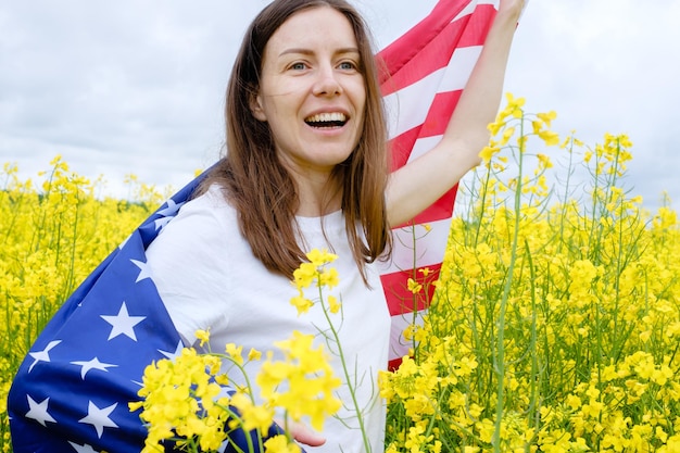 Young woman wrapping in American flag smiling widely among yellow flowers